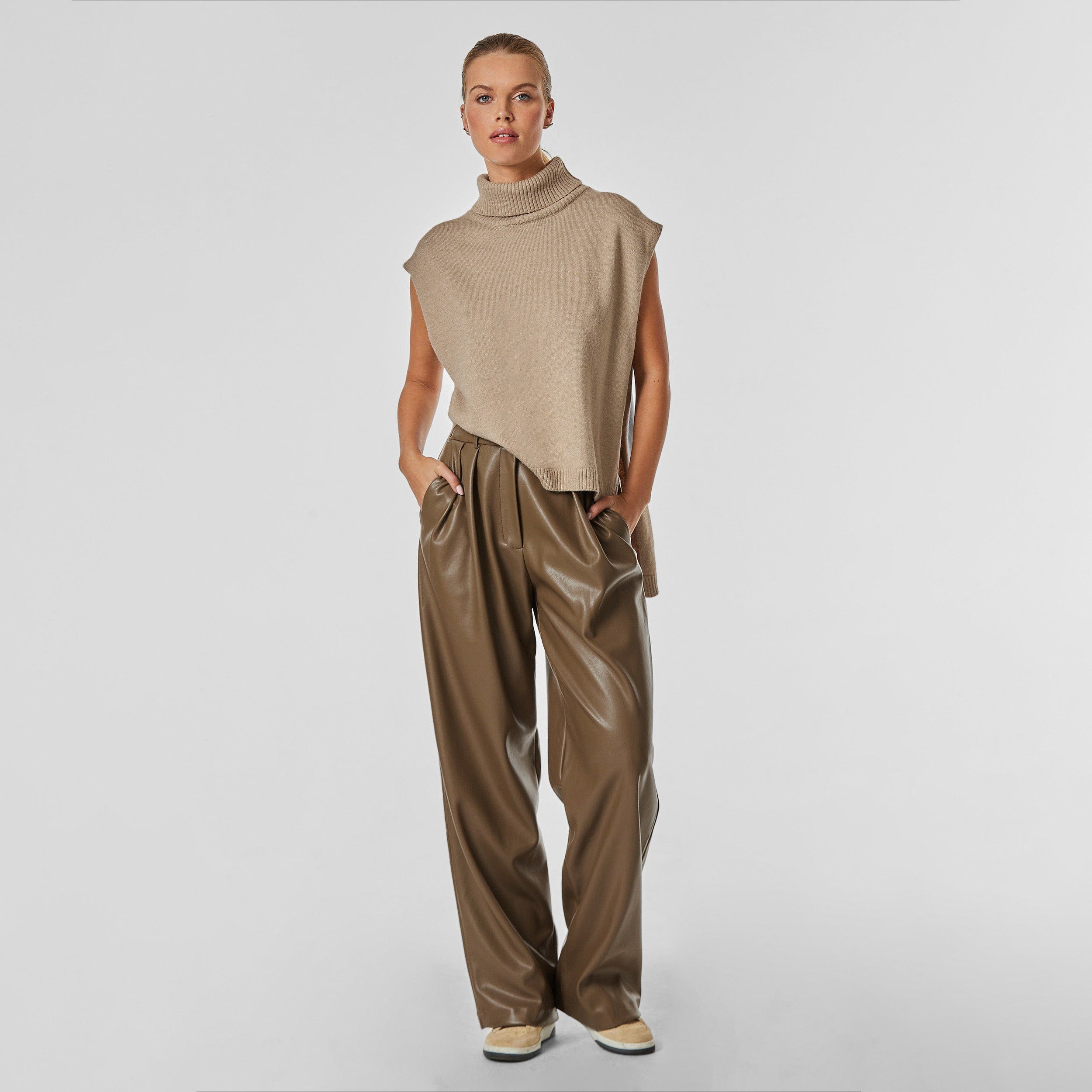 Full body front view of woman wearing sleeveless mocha sweater and brown high rise vegan leather pleated trousers.