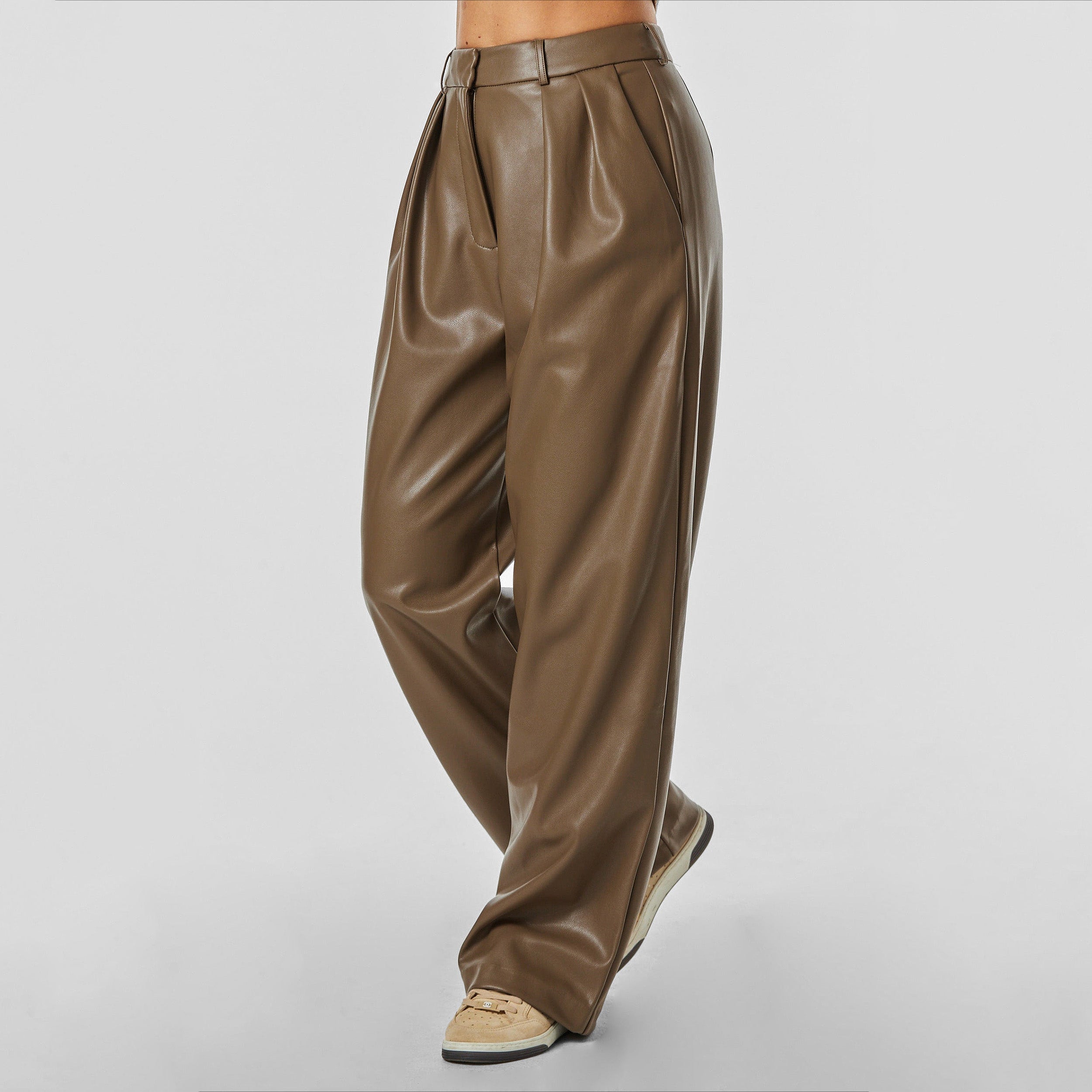 Side view of woman wearing brown high rise vegan leather pleated trousers.