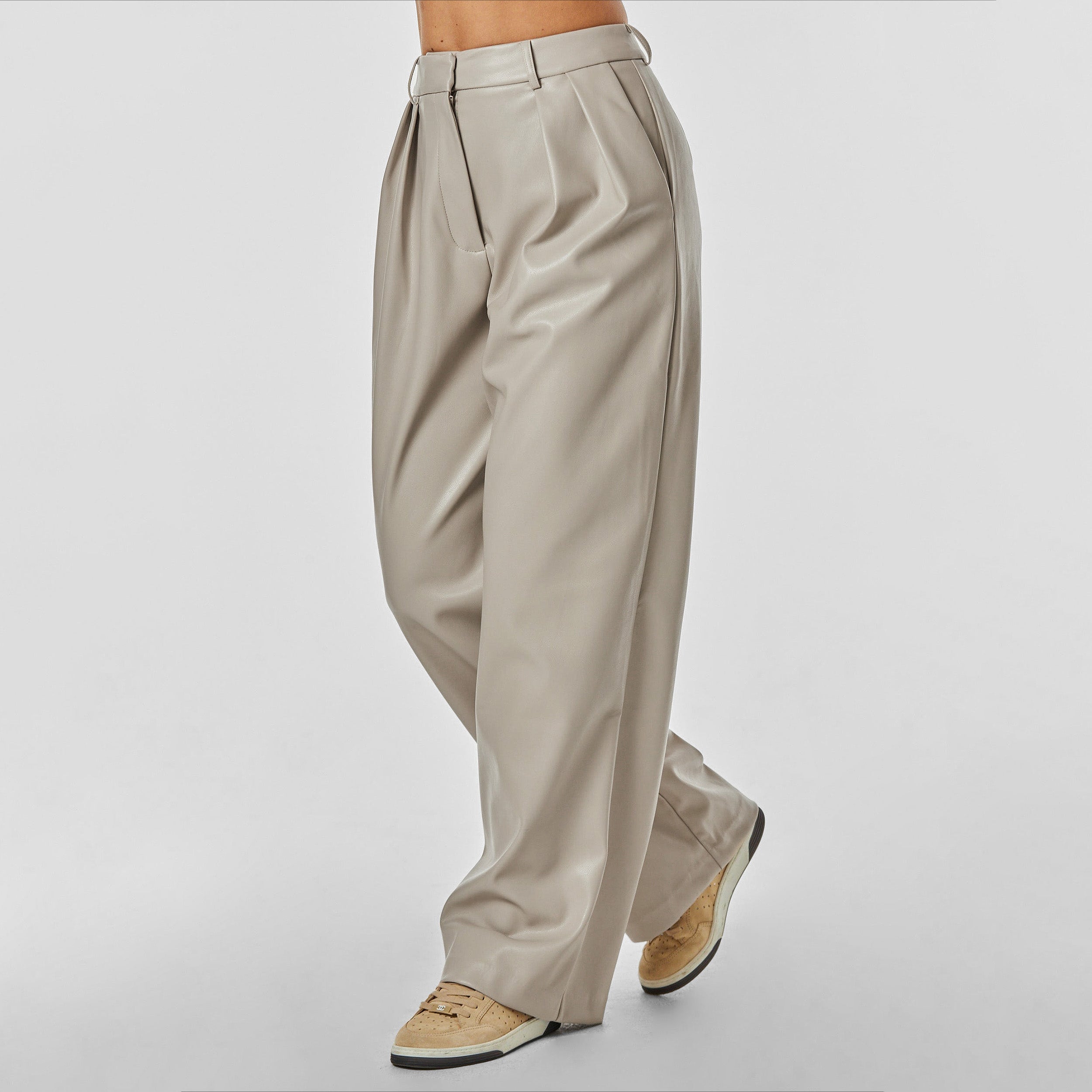 3/4 front view of woman wearing beige colored high rise vegan leather pleated trousers.