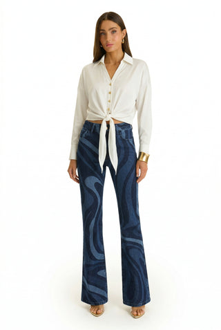 A woman faces forward wearing a white long-sleeve button-up shirt tied at the waist, high-rise Aria Flare Jean - Blue Wave, a gold cuff bracelet, and open-toe heels against a white background.