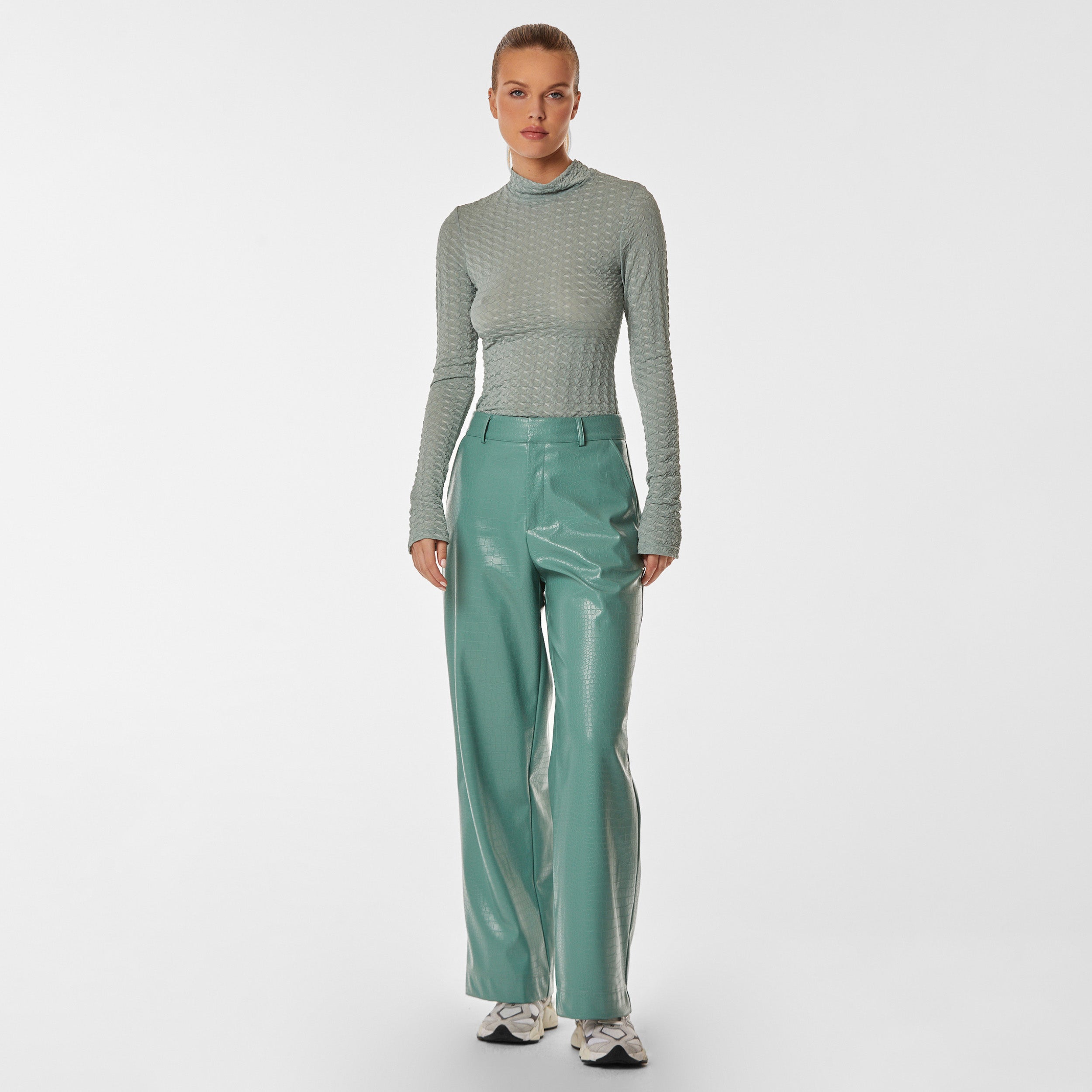 Fully body view of woman wearing green stretch mesh textured turtleneck sweater and mint green croco embossed faux leather pant.