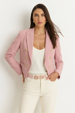 A woman with long brown hair and a neutral expression stands against a white background, wearing the Gabrielle Double Breasted Blazer in blush with gold buttons, paired with a white top and high-waisted cream pants with a pink belt.