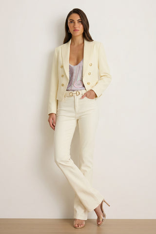A woman stands against a plain wall wearing the Gabrielle Double Breasted Blazer - Pearl, matching high-waisted pants, a patterned top, belt, and nude heels—effortlessly channeling quiet luxury with one hand in her pocket.
