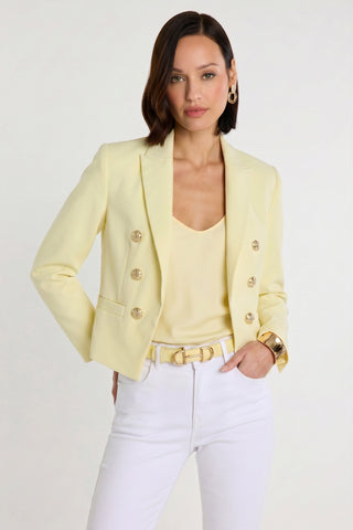 A woman models the Gabrielle Double Breasted Blazer in Sunshine Yellow with a matching light yellow top, white pants, and gold accessories, posing confidently with one hand in her pocket against a plain white background.