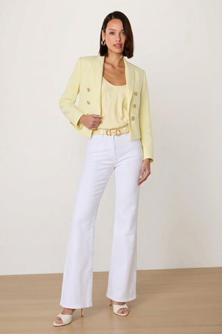 A woman stands indoors on a wooden floor wearing the Gabrielle Double Breasted Blazer in Sunshine Yellow, matching top, white high-waisted flared pants, yellow belt, white open-toe heels, and gold earrings. Her shoulder-length hair completes the look.