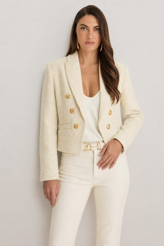A woman with long brown hair wears the Gabrielle Tweed Double Breasted Blazer in beige over a white top and high-waisted cream pants. She stands confidently against a light background, showcasing this chic blazer look.