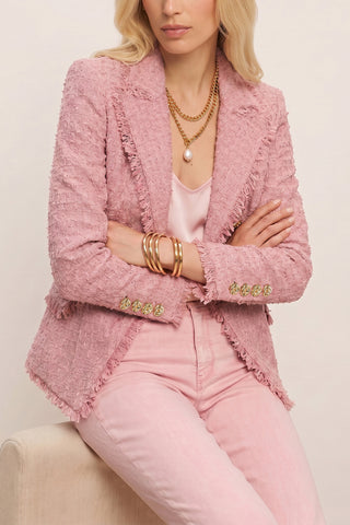 A woman sits with arms crossed, wearing the Giselle Single Breasted Tweed Blazer in blush over a light pink top and matching pants. She accessorizes with layered gold necklaces, a pearl pendant, and multiple gold bangles. Her head is partially out of frame.
