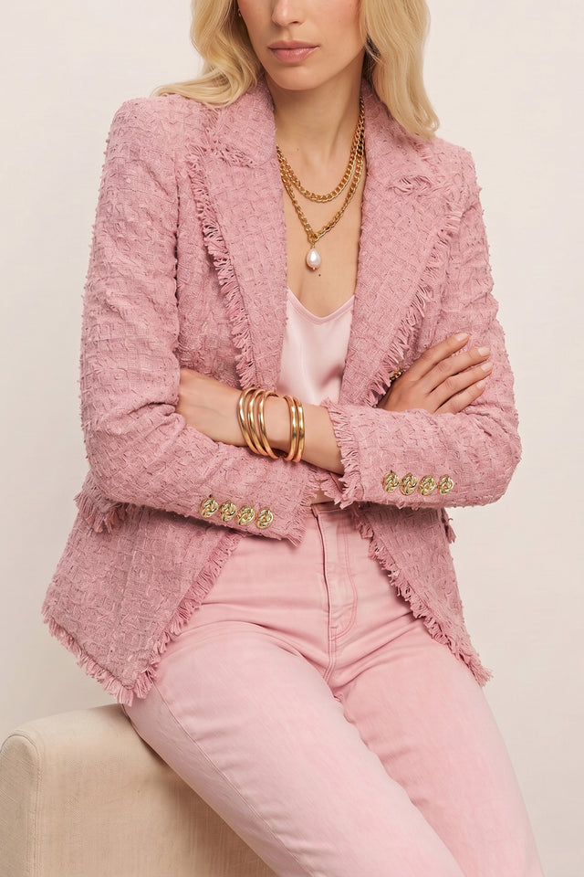 A woman sits with arms crossed, wearing the Giselle Single Breasted Tweed Blazer in blush over a light pink top and matching pants. She accessorizes with layered gold necklaces, a pearl pendant, and multiple gold bangles. Her head is partially out of frame.