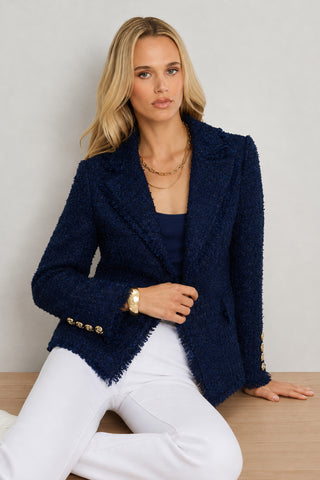 A woman with long blonde hair wears the Giselle Single Breasted Tweed Blazer in navy over a navy top and white pants. She is seated on a wooden surface against a light background, accessorized with a gold necklace and bracelet.