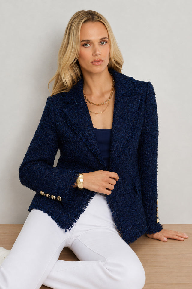 A woman with long blonde hair wears the Giselle Single Breasted Tweed Blazer in navy over a navy top and white pants. She is seated on a wooden surface against a light background, accessorized with a gold necklace and bracelet.
