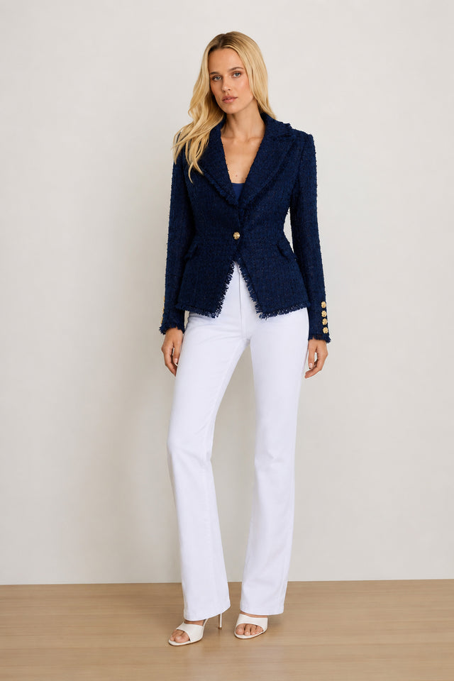 A woman stands on a wooden floor wearing the Giselle Single Breasted Tweed Blazer in navy with gold buttons, paired with white flared pants and open-toe heels against a plain, light background.