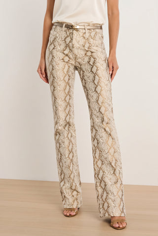 Wearing the Aria Flare Jean - Gold Python with a matching belt, white top, and open-toe heels, this look features four-way stretch denim for comfort and a chic snakeskin-inspired style. The outfit is set against a plain wall on a wooden floor.