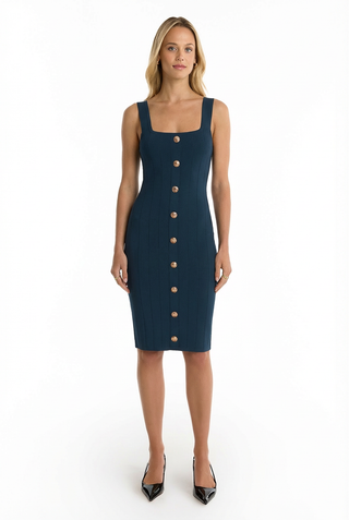 A woman stands facing forward in a fitted, sleeveless Helena Button Dress - Ocean with wide straps and bold gold buttons down the front, paired with black pointed-toe heels, against a white background.