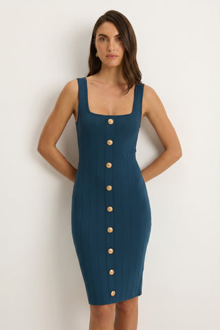 A woman stands against a plain light wall wearing the Helena Button Dress - Ocean, a fitted sleeveless knit midi dress with a square neckline and large round buttons down the front.