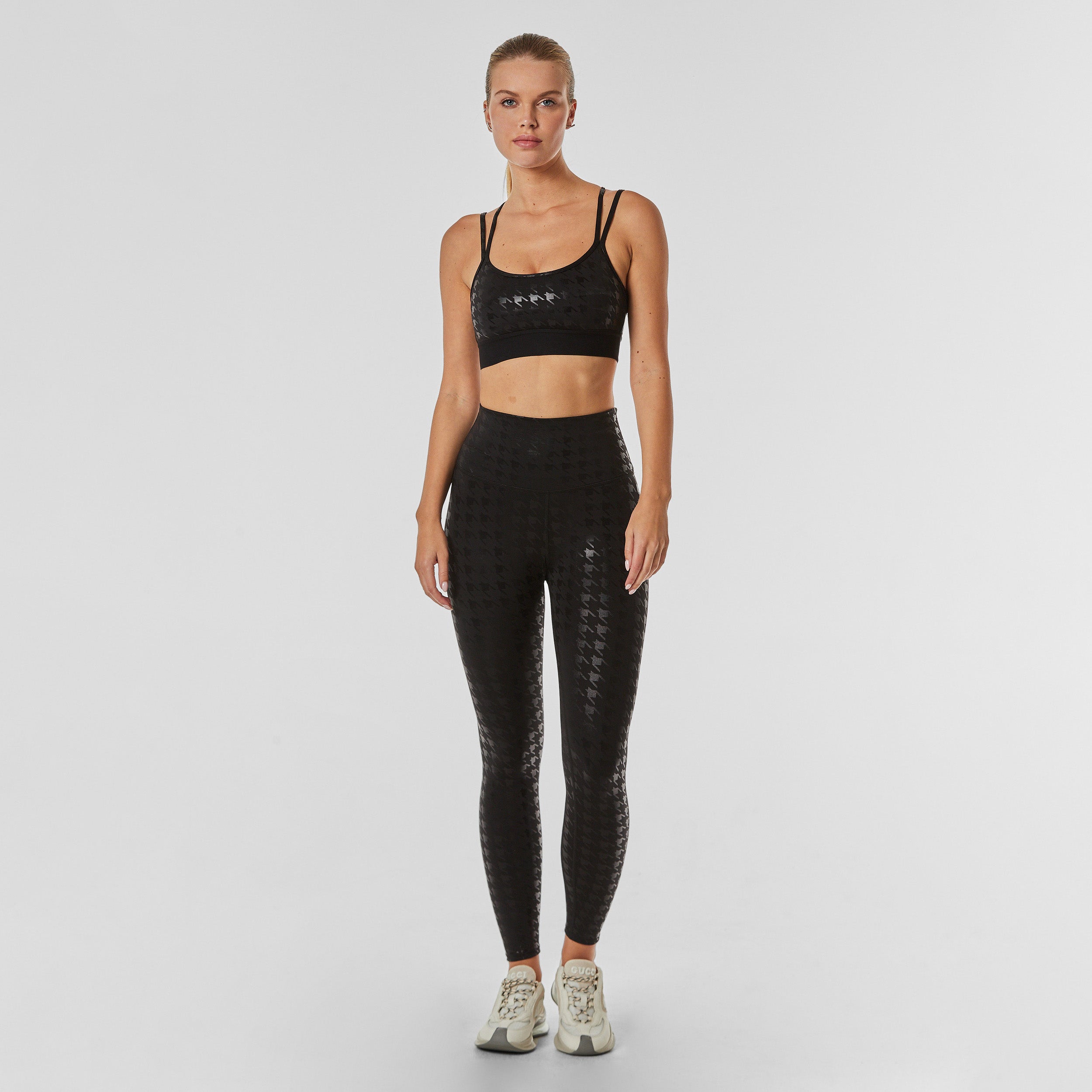 Full body view of woman wearing black on black glossy houndstooth patterned legging and matching bra