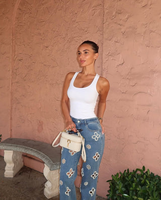 A woman with dark hair in a bun, wearing a white tank top and blue jeans with floral patterns, stands against a textured pink wall, holding a small cream-colored handbag by green plants and a stone bench.
