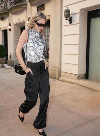 A woman wearing sunglasses, a sleeveless snakeskin print top, black wide-leg pants, and black heels carries a quilted black purse as she strolls confidently on a city sidewalk in front of an elegant building.