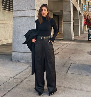 A woman stands on a city sidewalk wearing a black ribbed turtleneck sweater and wide-leg cargo pants with a belt, holding a black coat. Her pointed black shoes complete the look, set against tall buildings and leafy trees in the background.