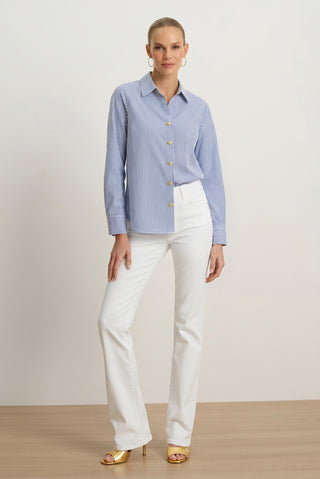 A woman stands indoors wearing the Jasmine Shirt in Blue Stripe paired with white pants and gold high-heeled sandals. Her hair is pulled back as she poses with one hand on her hip against a plain background, capturing effortless resort styling.