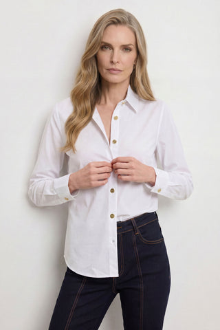 A woman with long, wavy blonde hair stands against a plain light background, looking at the camera and fastening her Jasmine Shirt - White with dark jeans.