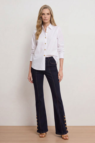A woman with long blond hair stands against a light background wearing the Jasmine Shirt in white and dark high-waisted flared jeans with gold ankle buttons, paired with tan heeled sandals.