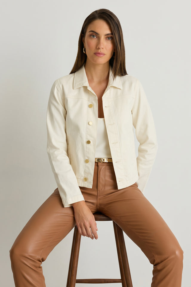 A woman with straight brown hair sits on a wooden stool, wearing the Joelle Jean Jacket in Ivory over a white top and tan leather pants. She gazes confidently at the camera against a plain light background.