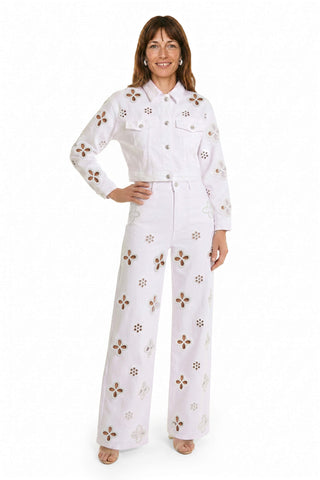 A woman smiles confidently in a white denim jacket and Kaia Pearl High Rise Jean - White, both featuring large cut-out flower designs and pearl accents, posing with one hand on her hip against a plain white background.