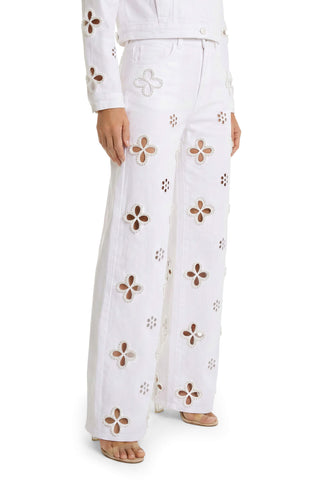 A person stands barefoot, wearing the Kaia Pearl High Rise Jean in white, featuring wide legs, large floral cutout designs, and subtle pearl embellishments. Matching cutouts are visible on the jacket sleeves. Only the lower body is shown.