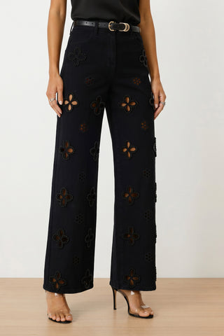 A person wearing the Kaia Pearl High Rise Jean - Black, featuring large floral cutouts with stud and pearl details, styled with a black belt, sleeveless black top, rings, and clear high-heeled sandals.