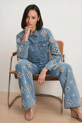 A woman with a bob haircut sits on a modern chair, wearing the Kaia Pearl High Rise Jean in Medium Blue and a matching denim jacket with white flower appliqués. She rests her chin on her hand, looking at the camera.