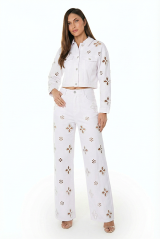 A woman models the Kaia Pearl High Rise Jean in white, featuring brown and tan floral cutouts. She pairs it with a matching cropped jacket, has long brown hair, and wears open-toe heels.