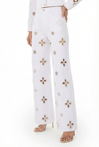 Wearing the Kaia Pearl High Rise Jean in white with wide legs and bold floral cutouts, paired with open-toe high-heeled sandals and a coordinating white jacket featuring matching floral accents.