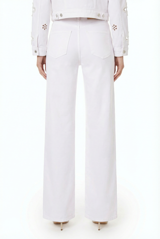 A person stands facing away, wearing the Kaia Pearl High Rise Jean in white—stretch denim wide-leg pants with two back pockets—paired with a white jacket featuring floral sleeve cutouts and heeled shoes.