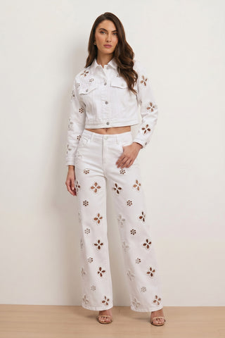 Woman wearing Kaia Pearl High Rise Jean in white—high-waisted wide-leg pants with floral cutout detail—paired with a cropped jacket, standing against a light background, long wavy hair and neutral sandals.