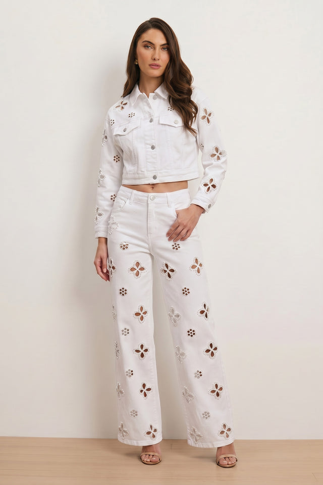 Woman wearing Kaia Pearl High Rise Jean in white—high-waisted wide-leg pants with floral cutout detail—paired with a cropped jacket, standing against a light background, long wavy hair and neutral sandals.