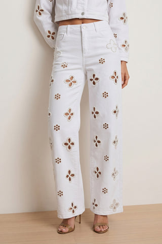 Wearing the Kaia Pearl High Rise Jean in white, featuring cut-out floral patterns, paired with a matching white jacket and gold open-toe heels, standing against a neutral background.