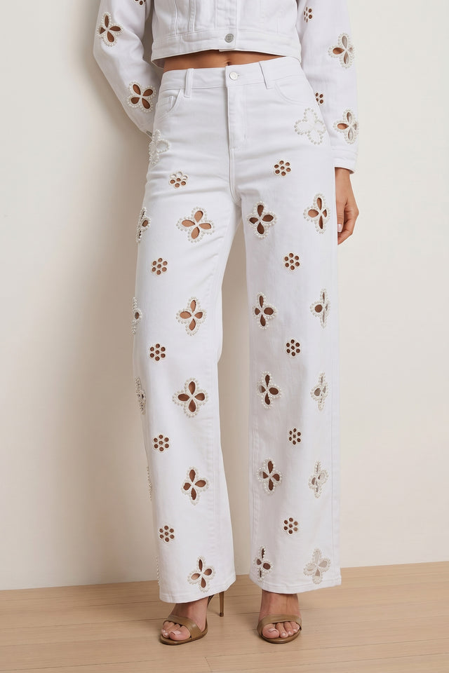 Wearing the Kaia Pearl High Rise Jean in white, featuring cut-out floral patterns, paired with a matching white jacket and gold open-toe heels, standing against a neutral background.