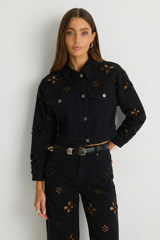 A woman with long brown hair wears the Kaia Pearl Jean Jacket in black, featuring floral cut-outs, paired with matching pants. She stands against a white wall, looking at the camera with a neutral expression.