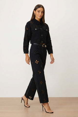 A woman wearing the Kaia Pearl Jean Jacket - Black, matching pants, a black belt, and black strappy heels walks on a light wooden floor against a plain white background.