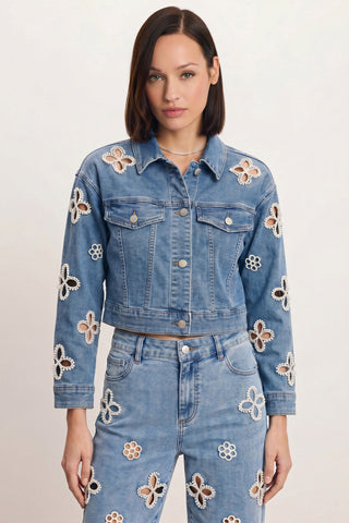 A woman with straight brown hair stands against a light background wearing the Kaia Pearl Jean Jacket in medium blue, paired with matching jeans featuring white and beige floral cutout patterns.