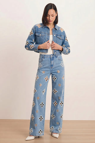 A person wears the Kaia Pearl Jean Jacket in medium blue with cream and black floral cutouts, paired with matching wide-leg jeans, a white top, and pointed white shoes against a plain light background.