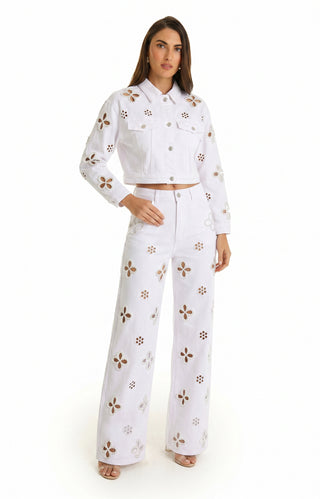 A woman with straight brown hair models the Kaia Pearl Jean Jacket in white, featuring cut-out floral patterns. She pairs it with matching high-waisted pants and open-toe shoes, posed against a white background.
