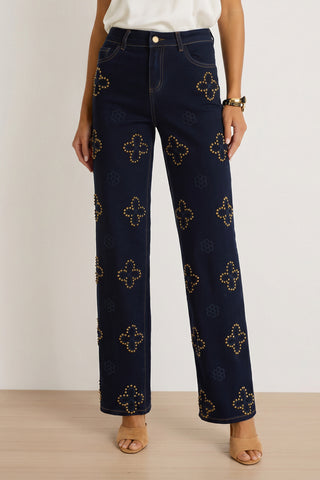 A person wearing the Kaia Studded High Rise Jean in indigo, paired with a white top, tan heels, and a gold bracelet, stands on a light wood floor against a white background.