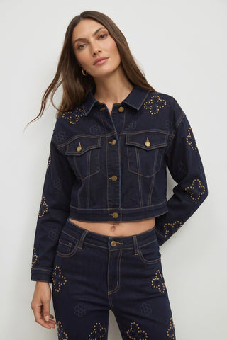 A woman wearing the Kaia Studded Jacket in Indigo—a dark blue cropped denim jacket featuring gold stud embellishments in floral and geometric patterns—paired with matching pants, stands against a plain light background.