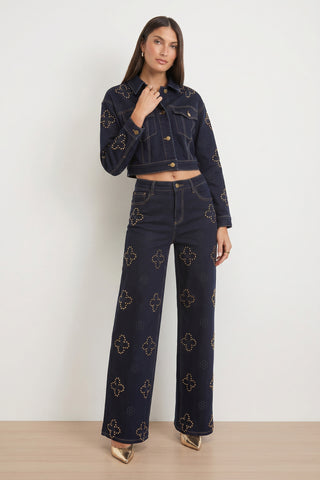 A woman with long straight hair and a neutral expression stands against a white wall wearing the Kaia Studded High Rise Jean in indigo, styled with a dark cropped denim jacket featuring gold embroidery and metallic pointed-toe heels.