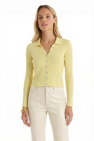 A woman with long blonde hair wears the Karolina Polo Shirt in Sunshine Yellow and high-waisted light beige pants, accessorized with gold hoop earrings and bracelets. Her arms are relaxed by her sides.