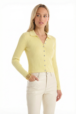 A woman with light hair wears the Karolina Polo Shirt in Sunshine Yellow and cream pants, standing with one hand in her pocket against a white background.
