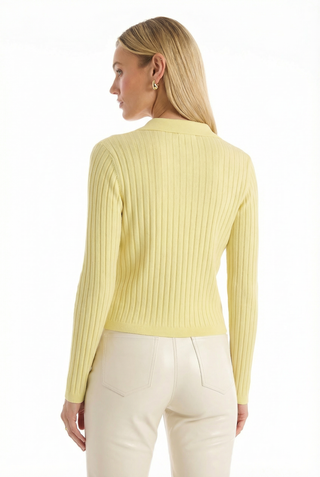 A woman with long blonde hair stands facing away against a white background, wearing the Karolina Polo Shirt in Sunshine Yellow and high-waisted cream pants.