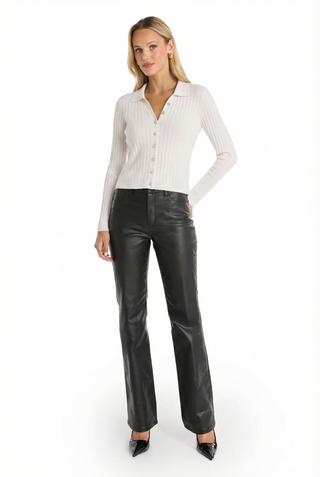 A woman with long blonde hair poses against a white background in the Karolina Polo Shirt - White, styled with black leather pants and pointed-toe heels. She stands confidently with one hand on her hip.