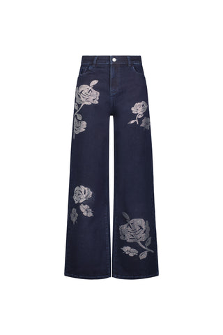The Kate Crystal Rose Jean - Indigo features a wide-leg silhouette with white floral embroidery and large crystal rose accents on the thighs and lower legs. High-waisted with front button closure, pockets, and a subtle four-way stretch for comfort.